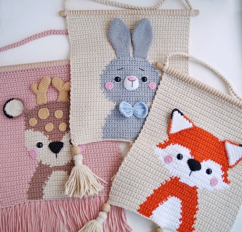 May include: Three crocheted wall hangings featuring animal designs. The first is a pink and brown deer with brown spots, the second is a beige bunny with a blue bow tie, and the third is a beige fox with orange and white fur.