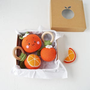 May include: A collection of orange crochet baby toys, including three round rattles with wooden rings and a slice, presented in a brown box. The toys are orange with white and green details, some with smiling faces.
