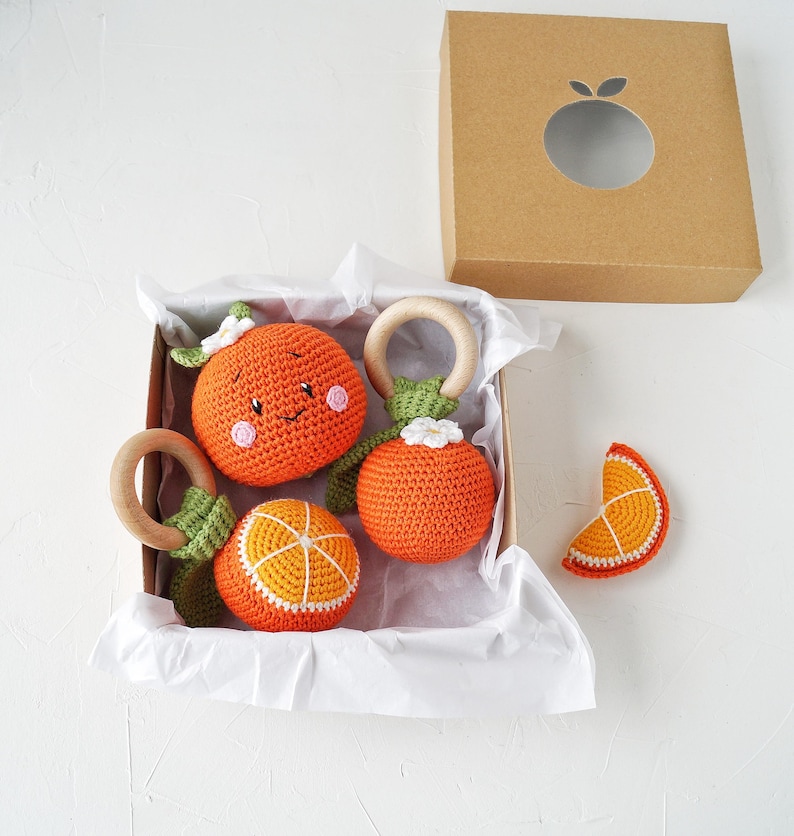 May include: A collection of orange crochet baby toys, including three round rattles with wooden rings and a slice, presented in a brown box. The toys are orange with white and green details, some with smiling faces.