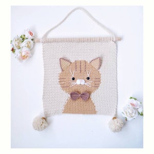 May include: A crocheted wall hanging featuring a brown cat with a bow tie. The cat is surrounded by a cream-colored crocheted background. The hanging has two white pom-poms on the bottom.