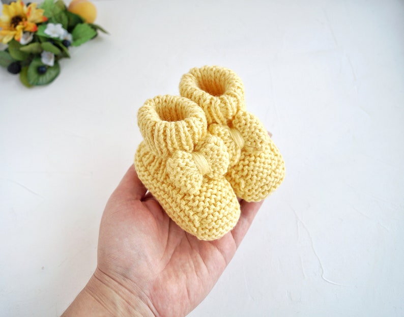 May include: A pair of yellow knitted baby booties with a bow detail. The booties are soft and cosy, perfect for keeping little feet warm.