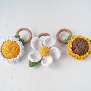 May include: Three crocheted flower teethers with wooden rings. The teethers are a yellow daisy, a white daisy, and a brown sunflower.