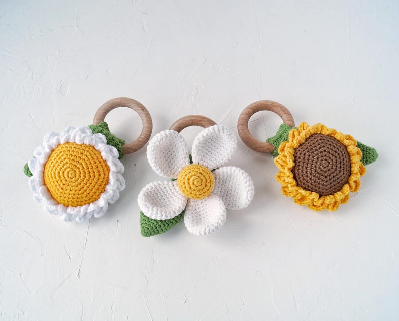 May include: Three crocheted flower teethers with wooden rings. The teethers are a yellow daisy, a white daisy, and a brown sunflower.