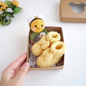 May include: A brown gift box with a white cutout of a bear's head. Inside the box are two yellow knitted baby booties and a crocheted bee rattle.