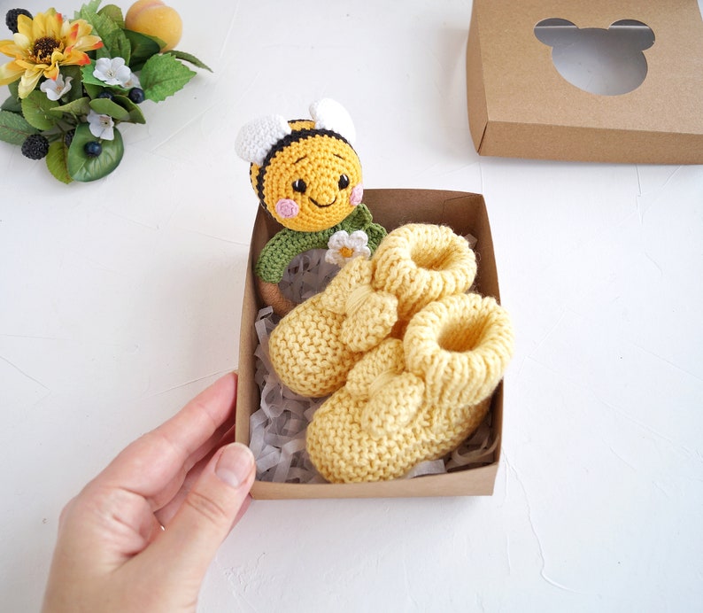 May include: A brown gift box with a white cutout of a bear's head. Inside the box are two yellow knitted baby booties and a crocheted bee rattle.