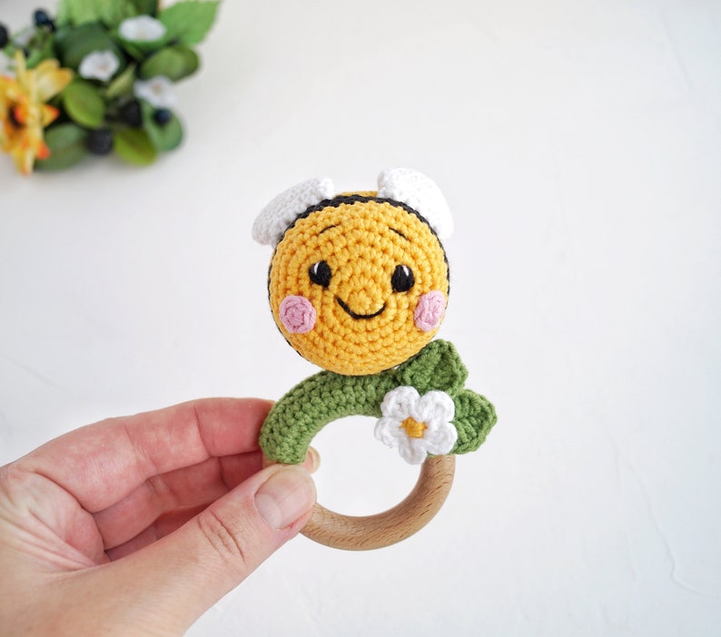 May include: A crocheted bee rattle toy with a wooden ring. The bee is yellow and black with a smiling face and pink cheeks. The bee is attached to a green crocheted flower with a white centre.