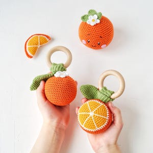 May include: A collection of crocheted orange-themed baby toys. The toys include a slice, a whole orange with a face, and two oranges with wooden rings. The oranges are orange with white and green accents. Handmade.