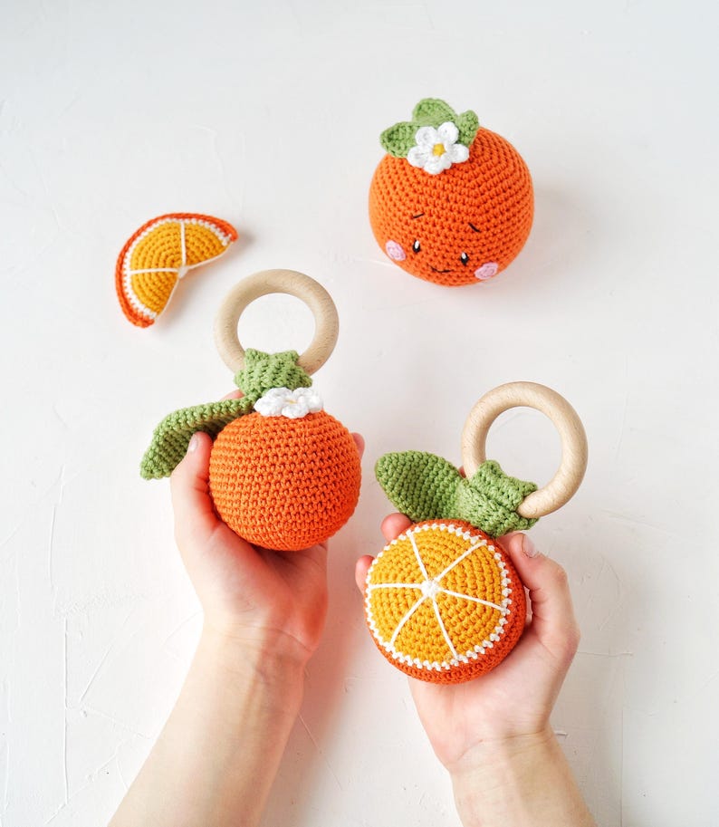 May include: A collection of crocheted orange-themed baby toys. The toys include a slice, a whole orange with a face, and two oranges with wooden rings. The oranges are orange with white and green accents. Handmade.