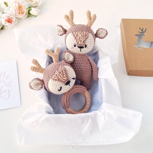 Peut inclure: Deux jouets en forme de cerf crochet&eacute;s, dont un hochet, dans une bo&icirc;te doubl&eacute;e de papier blanc. Les cerfs sont bruns et cr&egrave;me, avec des &eacute;l&eacute;ments en bois. Une bo&icirc;te en papier kraft avec un cerf d&eacute;coup&eacute; et une carte "Hello Baby" sont &eacute;galement pr&eacute;sents.