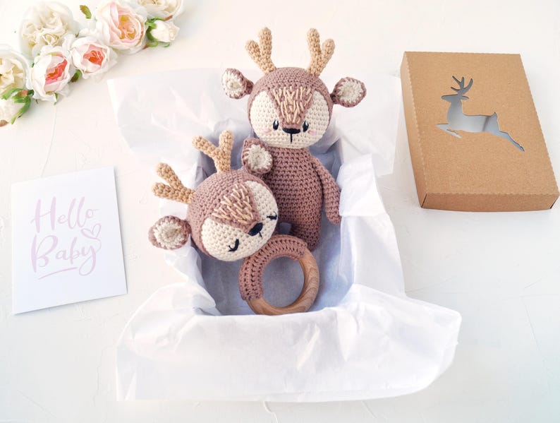Peut inclure: Deux jouets en forme de cerf crochet&eacute;s, dont un hochet, dans une bo&icirc;te doubl&eacute;e de papier blanc. Les cerfs sont bruns et cr&egrave;me, avec des &eacute;l&eacute;ments en bois. Une bo&icirc;te en papier kraft avec un cerf d&eacute;coup&eacute; et une carte "Hello Baby" sont &eacute;galement pr&eacute;sents.