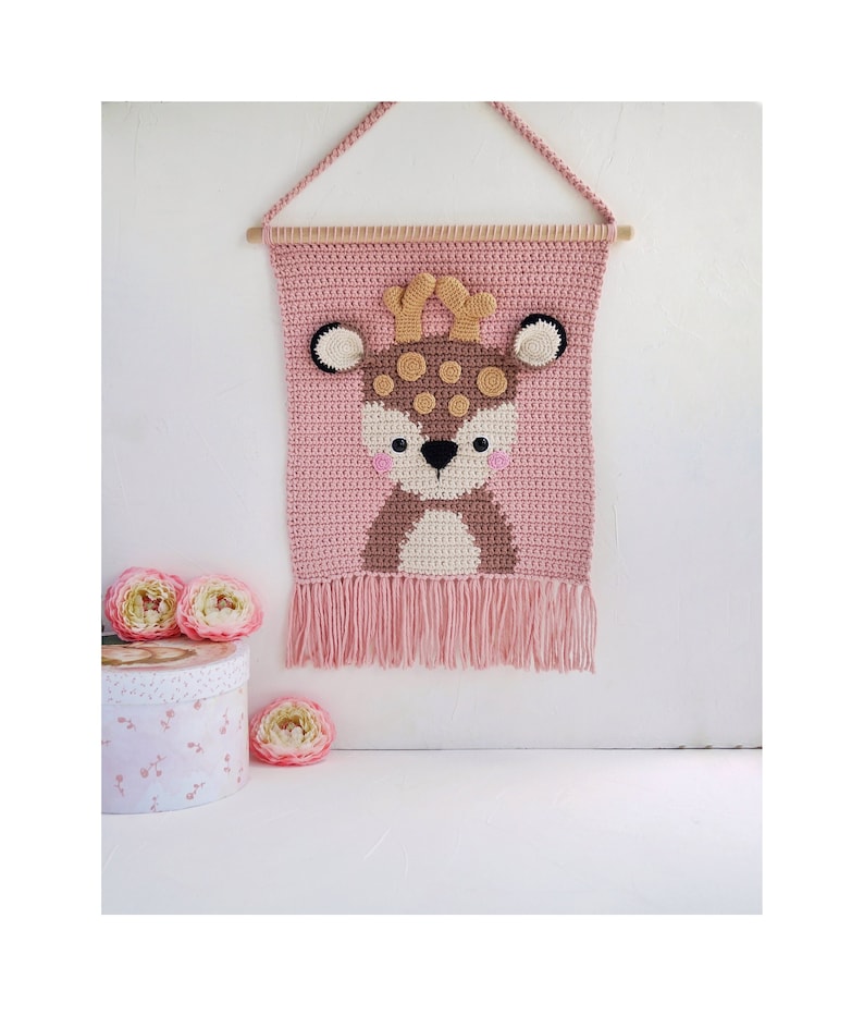 May include: A pink crocheted wall hanging with a brown deer face in the center. The deer has brown antlers, brown spots, and a white nose. The hanging has a fringe at the bottom.