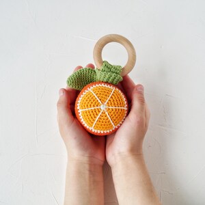 May include: A crocheted orange slice baby toy with a wooden ring and green leaves. The orange slice is orange with white and red details. The toy is designed for babies and toddlers.