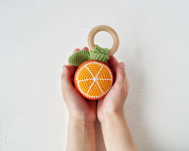 May include: A crocheted orange slice baby toy with a wooden ring and green leaves. The orange slice is orange with white and red details. The toy is designed for babies and toddlers.