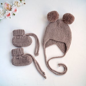 K&ouml;nnte beinhalten: Ein Set aus gestrickten Babyschuhen und einer passenden M&uuml;tze mit Bommeln. Die Schuhe und die M&uuml;tze sind hellbraun.