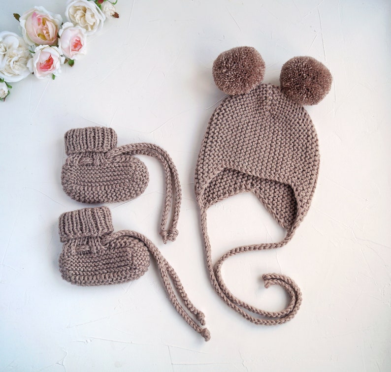 K&ouml;nnte beinhalten: Ein Set aus gestrickten Babyschuhen und einer passenden M&uuml;tze mit Bommeln. Die Schuhe und die M&uuml;tze sind hellbraun.