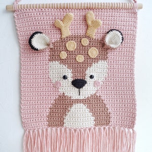 May include: A pink crocheted wall hanging with a brown deer face. The deer has brown antlers, brown eyes, and a white nose. The wall hanging has a fringe at the bottom.