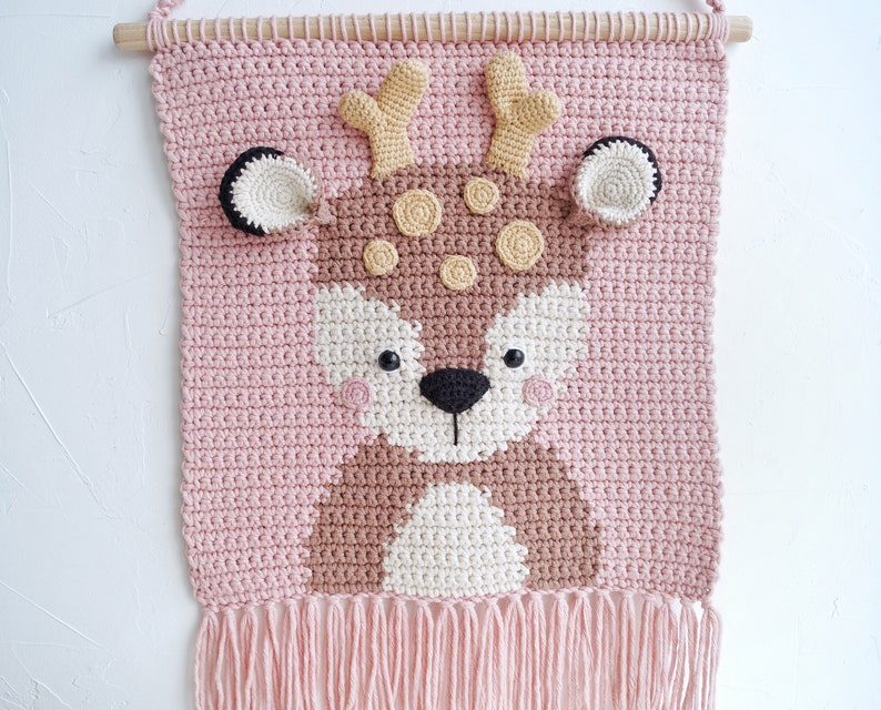 May include: A pink crocheted wall hanging with a brown deer face. The deer has brown antlers, brown eyes, and a white nose. The wall hanging has a fringe at the bottom.