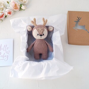 K&ouml;nnte beinhalten: Ein braunes, geh&auml;keltes Stofftier in Form eines Rehs mit wei&szlig;em Gesicht und Geweih befindet sich in einer wei&szlig;en Geschenkbox aus Seidenpapier. Links im Bild ist eine Karte mit dem Text "Hello Baby" zu sehen.