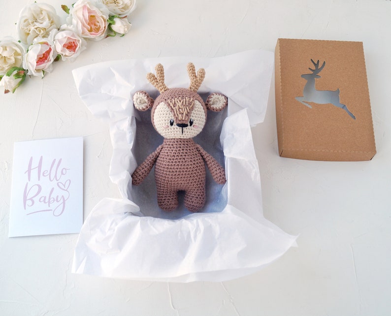 K&ouml;nnte beinhalten: Ein braunes, geh&auml;keltes Stofftier in Form eines Rehs mit wei&szlig;em Gesicht und Geweih befindet sich in einer wei&szlig;en Geschenkbox aus Seidenpapier. Links im Bild ist eine Karte mit dem Text "Hello Baby" zu sehen.