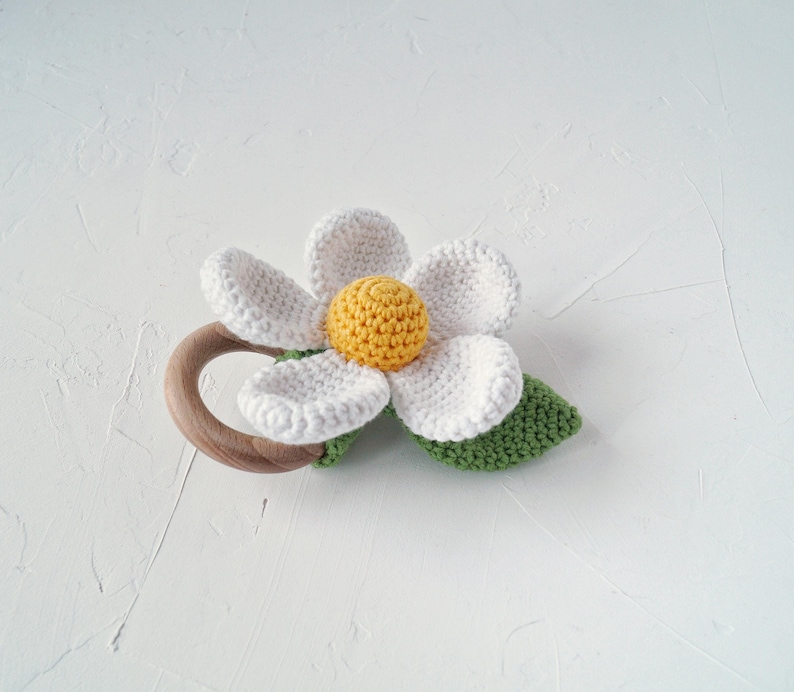 May include: A crocheted white flower with a yellow center attached to a wooden teething ring. The flower has green leaves.