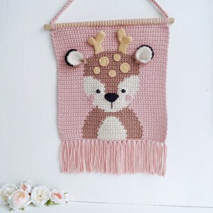 May include: A pink crocheted wall hanging with a brown deer face in the center. The deer has black eyes, a brown nose, and yellow antlers. The wall hanging has a fringe at the bottom.