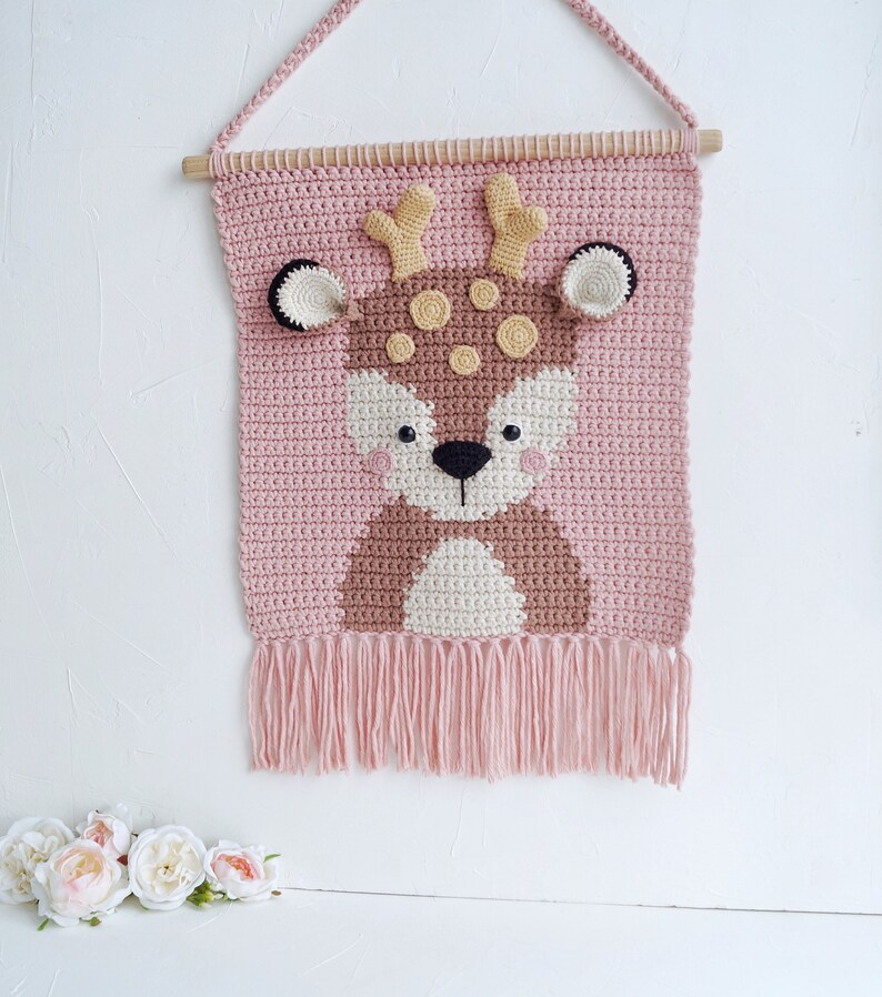 May include: A pink crocheted wall hanging with a brown deer face in the center. The deer has black eyes, a brown nose, and yellow antlers. The wall hanging has a fringe at the bottom.