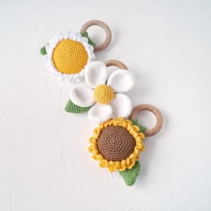 May include: Three crocheted flower teethers with wooden rings. The flowers are a yellow daisy, a white daisy, and a brown and yellow sunflower.