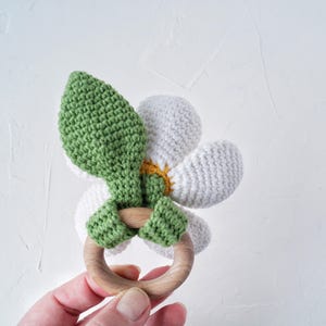 May include: A crocheted flower-shaped teether with a wooden ring. The flower is white with a yellow center and green leaves.