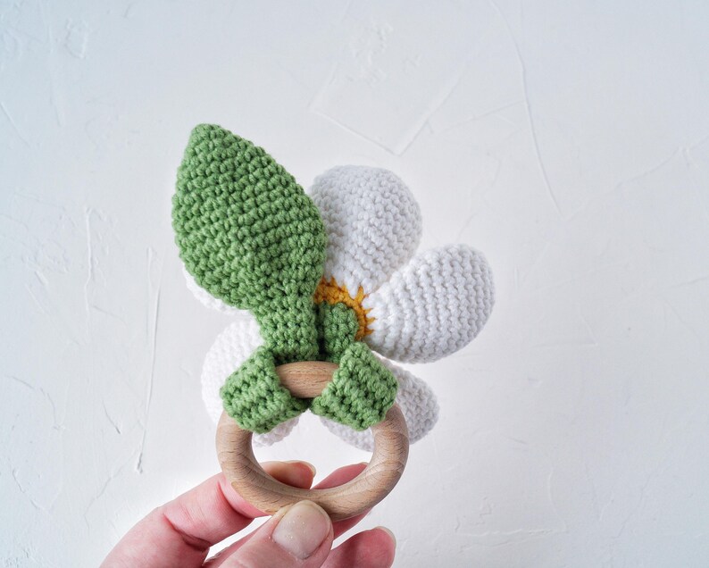 May include: A crocheted flower-shaped teether with a wooden ring. The flower is white with a yellow center and green leaves.