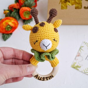 May include: A crocheted giraffe rattle with a wooden ring. The giraffe is yellow with brown spots and green fabric around its neck. The ring is engraved with the name "Austin".