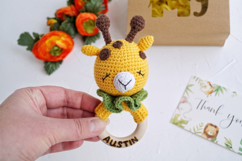 May include: A crocheted giraffe rattle with a wooden ring. The giraffe is yellow with brown spots and green fabric around its neck. The ring is engraved with the name "Austin".