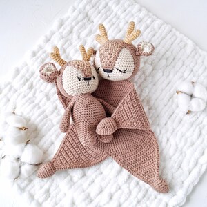 Peut inclure: Deux jouets en forme de cerf crochet&eacute;s, de couleur rose clair. L'un est une petite poup&eacute;e, l'autre une grande couverture de s&eacute;curit&eacute; avec une t&ecirc;te de cerf. Les cerfs ont des bois beiges, des visages cr&egrave;me et les yeux ferm&eacute;s, sugg&eacute;rant le sommeil. Les jouets sont sur une couverture blanche textur&eacute;e.