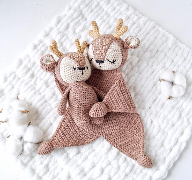 Peut inclure: Deux jouets en forme de cerf crochet&eacute;s, de couleur rose clair. L'un est une petite poup&eacute;e, l'autre une grande couverture de s&eacute;curit&eacute; avec une t&ecirc;te de cerf. Les cerfs ont des bois beiges, des visages cr&egrave;me et les yeux ferm&eacute;s, sugg&eacute;rant le sommeil. Les jouets sont sur une couverture blanche textur&eacute;e.
