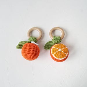 May include: Two crocheted orange-shaped baby toys with wooden rings. One orange is whole, the other is sliced, revealing a yellow and white interior. Both have green leaves and white accents.