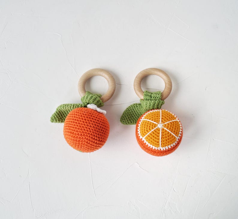 May include: Two crocheted orange-shaped baby toys with wooden rings. One orange is whole, the other is sliced, revealing a yellow and white interior. Both have green leaves and white accents.