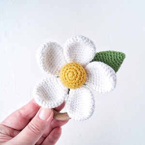 May include: A crocheted white flower with a yellow center and a green leaf attached to a wooden ring. The flower is a baby toy.