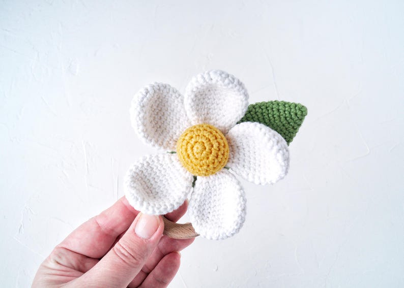 May include: A crocheted white flower with a yellow center and a green leaf attached to a wooden ring. The flower is a baby toy.