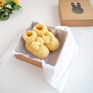 May include: A pair of yellow knitted baby booties with a bow detail, presented in a brown gift box with white tissue paper.
