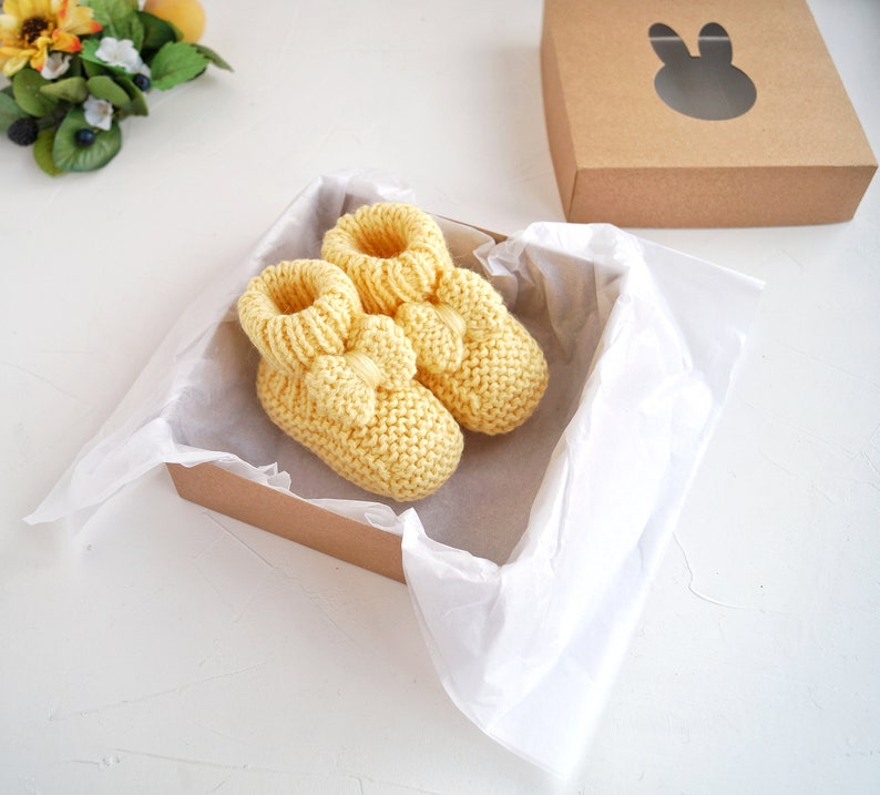 May include: A pair of yellow knitted baby booties with a bow detail, presented in a brown gift box with white tissue paper.
