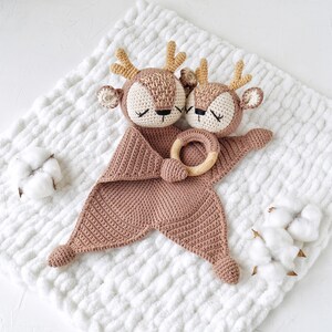 Peut inclure: Une couverture de s&eacute;curit&eacute; pour b&eacute;b&eacute; faite &agrave; la main au crochet, repr&eacute;sentant deux t&ecirc;tes de cerf aux yeux ferm&eacute;s, des bois brun clair et un anneau en bois. La couverture est de couleur rose clair et repose sur une surface blanche textur&eacute;e. Des cotons sont plac&eacute;s autour de la couverture.