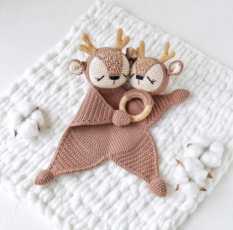 Peut inclure: Une couverture de s&eacute;curit&eacute; pour b&eacute;b&eacute; faite &agrave; la main au crochet, repr&eacute;sentant deux t&ecirc;tes de cerf aux yeux ferm&eacute;s, des bois brun clair et un anneau en bois. La couverture est de couleur rose clair et repose sur une surface blanche textur&eacute;e. Des cotons sont plac&eacute;s autour de la couverture.