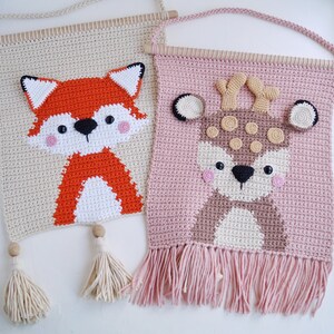 May include: Two crocheted wall hangings with animal designs. The first is a beige wall hanging with a red fox design. The second is a pink wall hanging with a brown deer design.