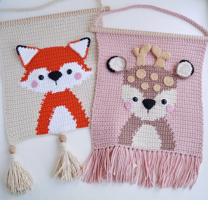 May include: Two crocheted wall hangings with animal designs. The first is a beige wall hanging with a red fox design. The second is a pink wall hanging with a brown deer design.