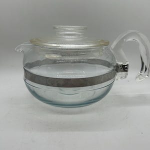 Puede incluir: Tetera de vidrio transparente con cuerpo redondeado, asa curva y boquilla. La tetera tiene una tapa de vidrio transparente y una banda metálica plateada en el centro. El diseño es sencillo y clásico.