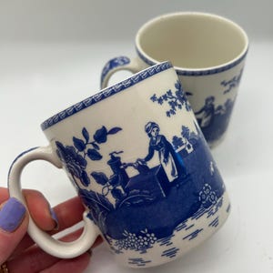 Könnte beinhalten: Zwei Keramikbecher mit blau-weißem Pastoraldesign. Die Becher haben einen cremefarbenen Boden mit blauen Henkeln und Rändern. Das blaue Design zeigt Blumen- und Landschaftsszenen. Ein Becher ist im Fokus, gehalten von einer Hand.