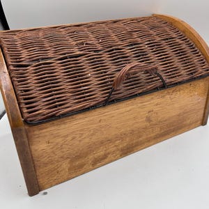 Op de afbeelding: Een rechthoekige brooddoos van hout met een geweven rieten deksel. De doos is gemaakt van lichtbruin hout met een donkerbruin geweven bovenkant. Een gebogen handvat is aan het deksel bevestigd.