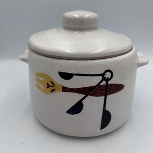 May include: A speckled cream ceramic jar with a lid and two handles. The jar features a whimsical design of a yellow broom and black and brown kitchen utensils. A charming addition to any kitchen.