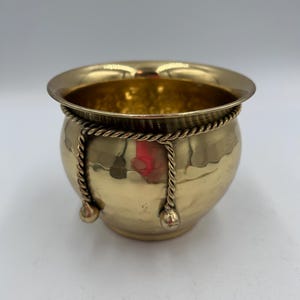 Puede incluir: Un cuenco pequeño y redondo de color latón con un borde ancho. El cuenco está adornado con un detalle similar a una cuerda alrededor del centro y dos borlas decorativas. El interior del cuenco también es de color latón. El cuenco tiene una superficie brillante y reflectante.