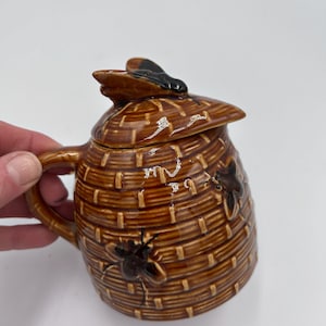 May include: A brown ceramic honey pot with a lid and handle, shaped like a woven beehive. The lid is topped with a black and brown bee figurine, and the pot features embossed bee details. The pot is held by a hand.