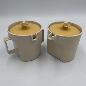 May include: Two vintage cream-colored plastic containers with mustard yellow lids. Each container has a handle and a spout. The lids have a small, round knob in the center. The containers are likely for sugar and creamer.